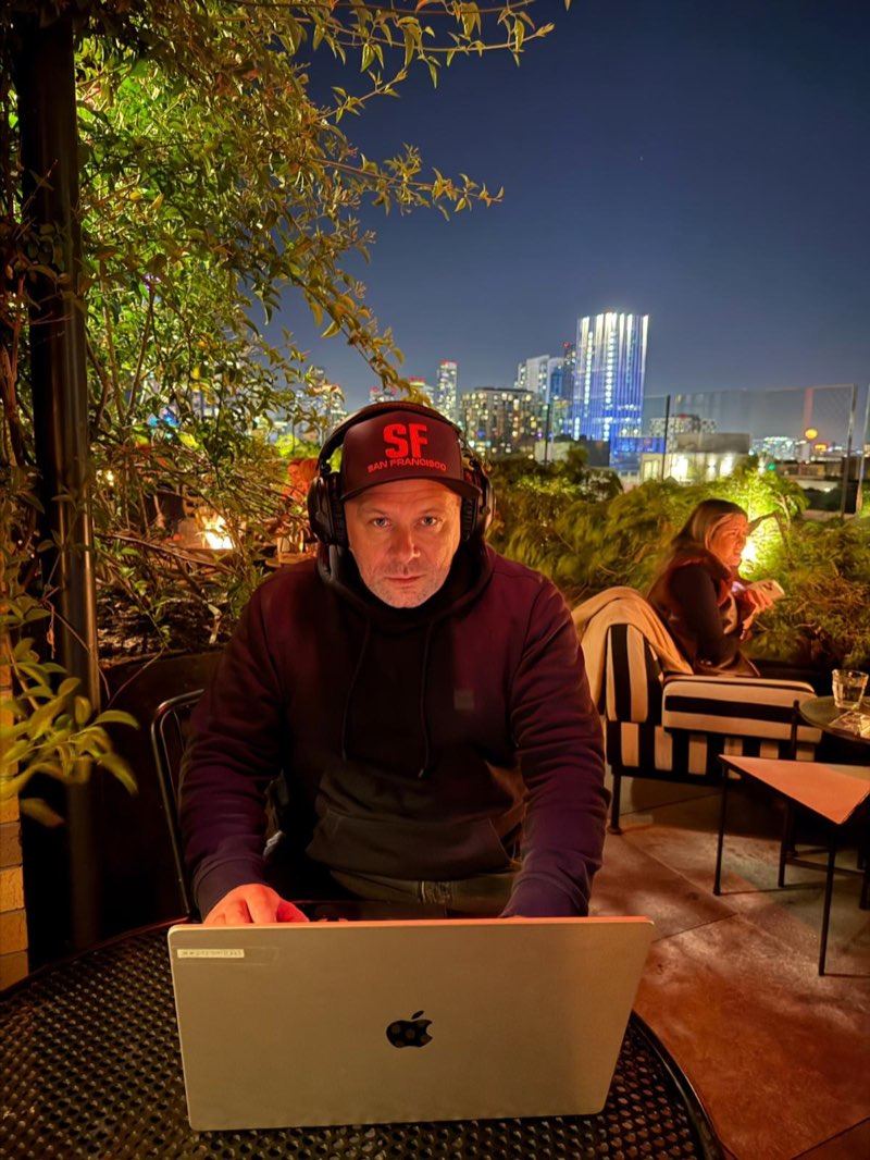 Constantin Crismaru working at a laptop on a rooftop terrace at night, with a lit city skyline behind him.